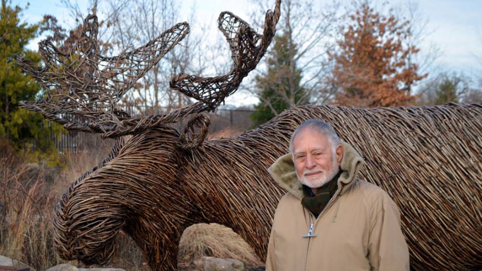 Robert Mark poses in front of a sculpture of a moose made of tree branches