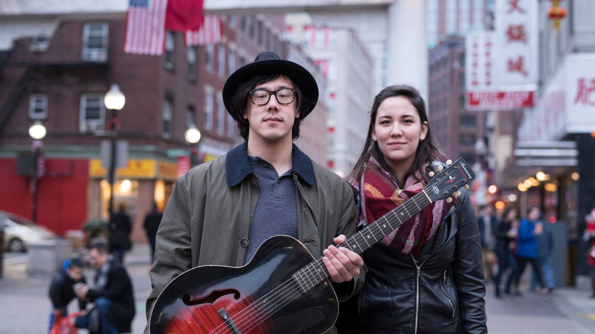 Young man in a hat holding an acoustic guitar standing next to a young woman