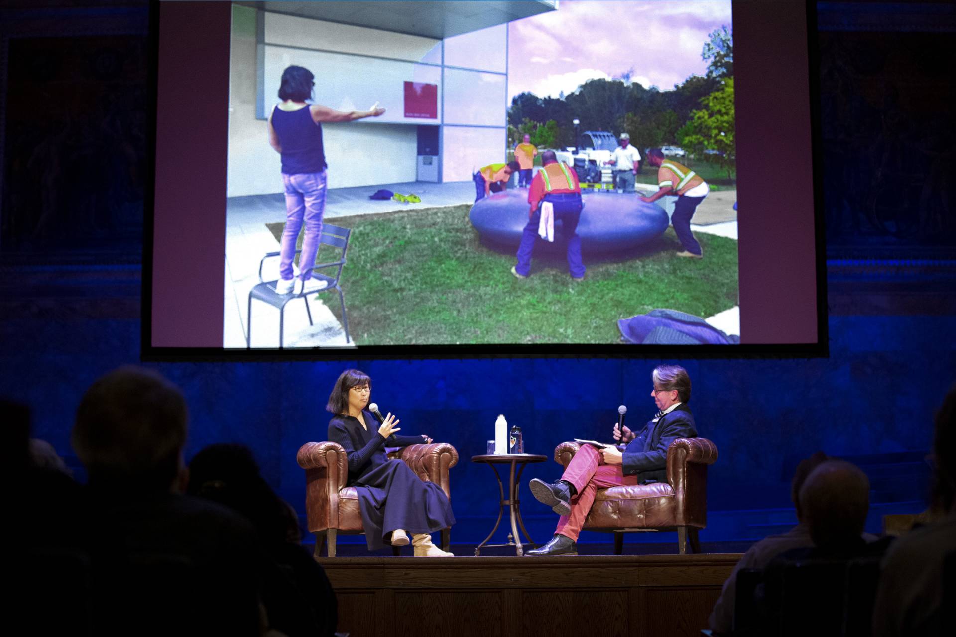 Maya Lin speaks with James Steward on stage about her installation and projects on a screen an image of her directing her colleagues positioning a large grey inner tube on a grassy square