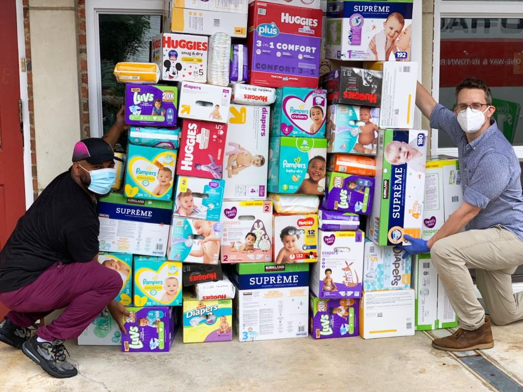 Two people wearing masks pose next to a collection of bulk diaper boxes