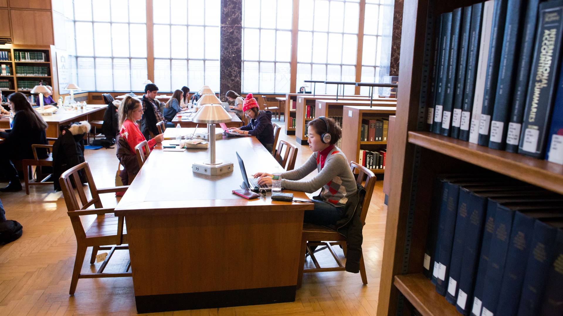 Students study at tables in firestone library in front of large windows