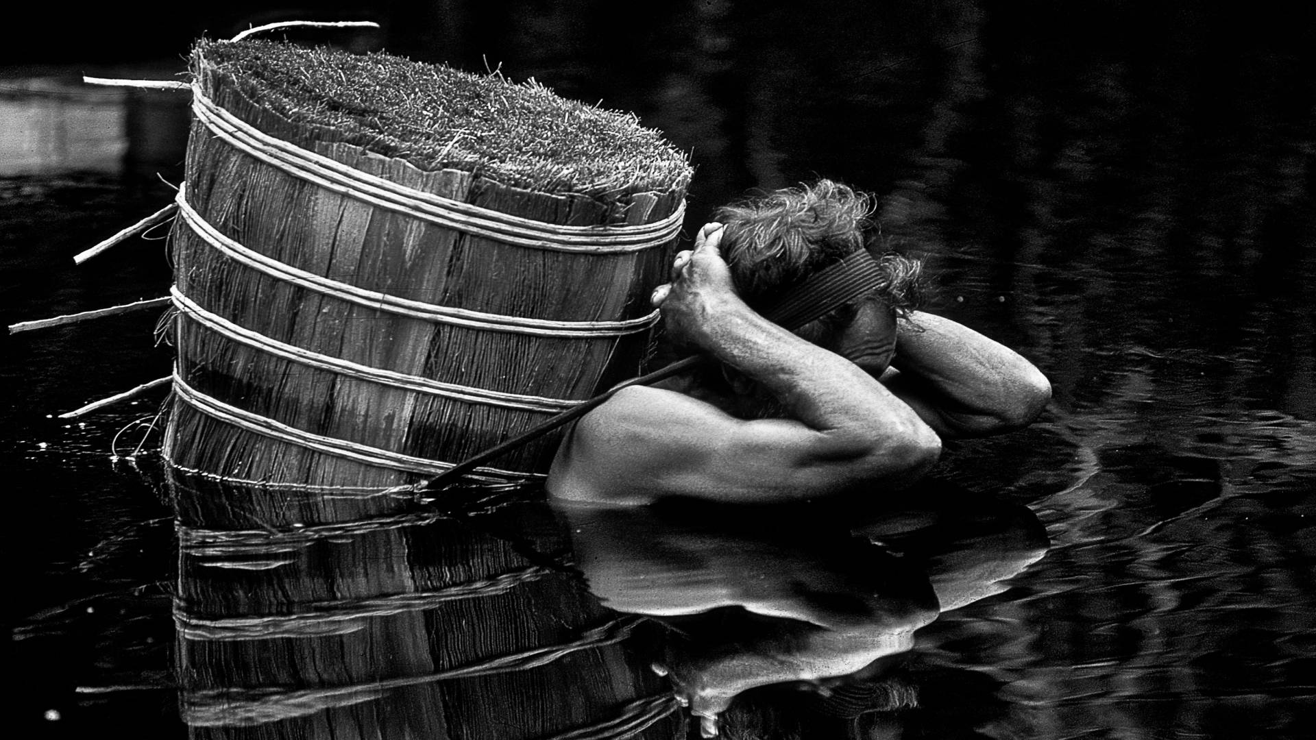 A man in water carying a barrel on his back