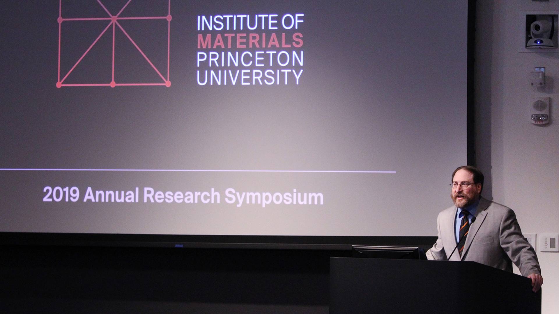 Craig Arnold standing at podium in front of screen with the words "Institute of Materials Princeton University 2019 Annual Research Symposium"