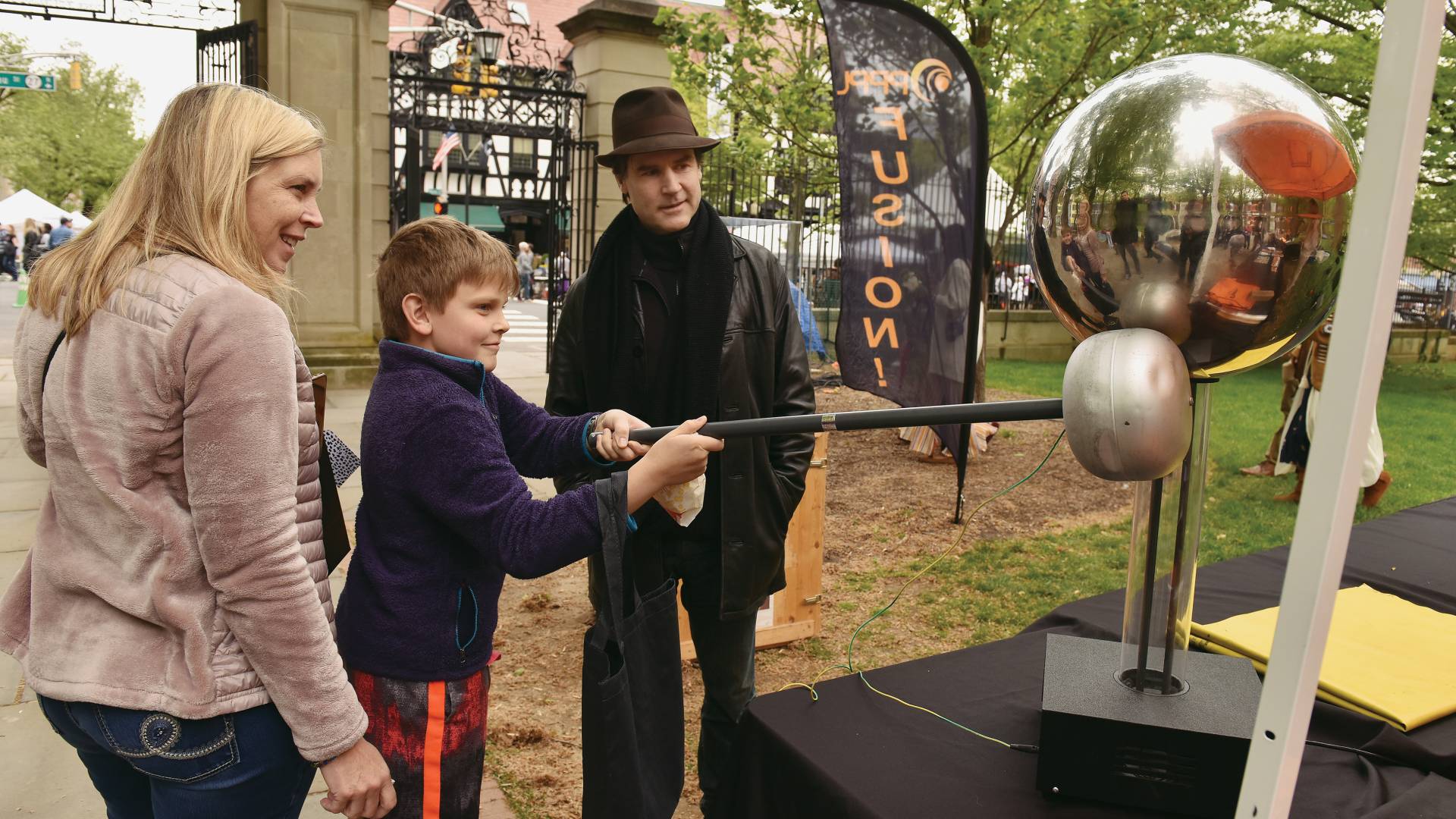 A boy holds a stick with a silver colored ball at the end close to to base of a silver sphere while his mother and a man watch on