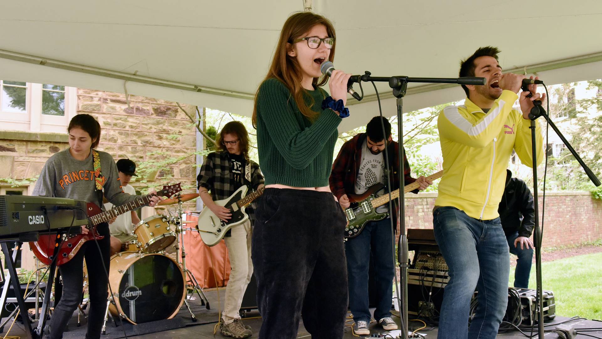 Young woman and a young man sing into microphones while a rock band play behind them