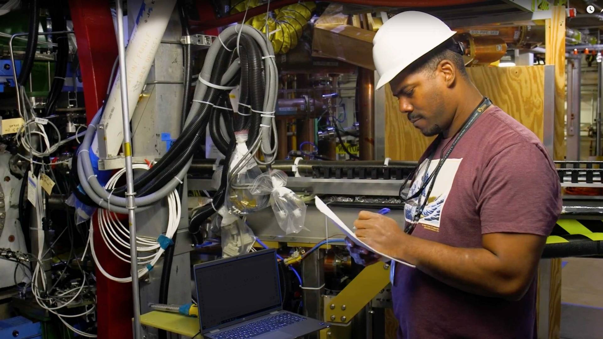 A man in a hard hat writes on papers in a machine room with many pipes