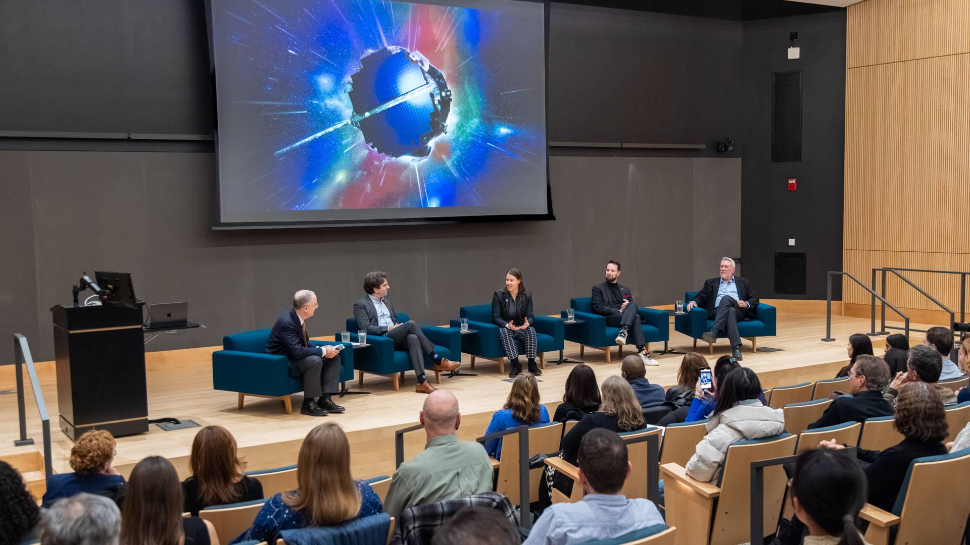 Panelists sit in front of a big screen that shows IMAP, a circular spacecraft covered in solar panels