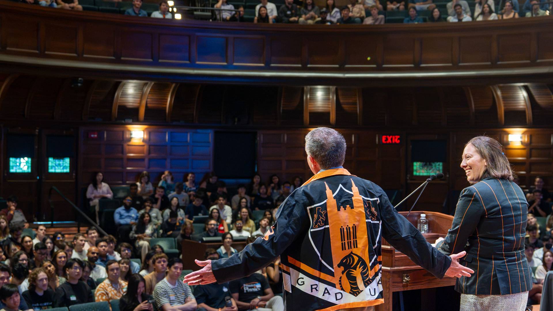 An alumus in a grad school class jacket speaks from the stage at Richardson.