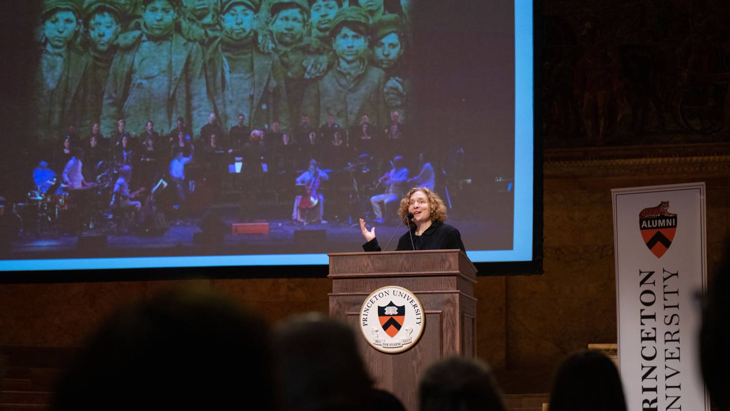 Julia Wolfe speaks at the podium in Richardson Hall, with a projection of street urchins behind 