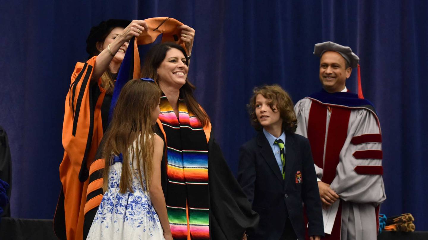 A graduate student having a hood placed over her head as her children look on. Livestream of ceremony