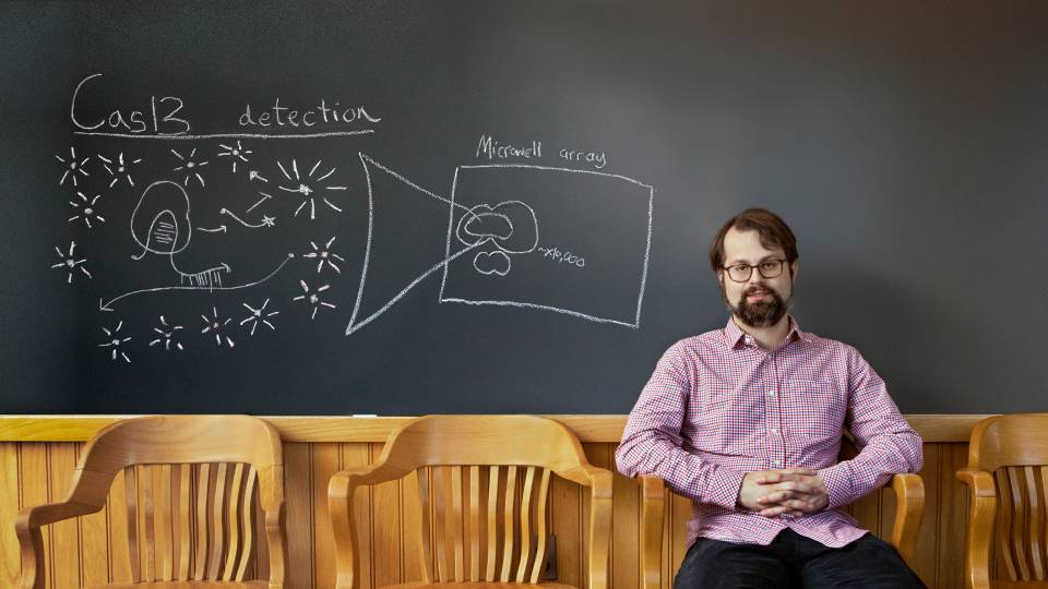 Man in front of a blackboard that has "Cas-13 detection" with star-like shapes underneath and pointing to "Microwell array"