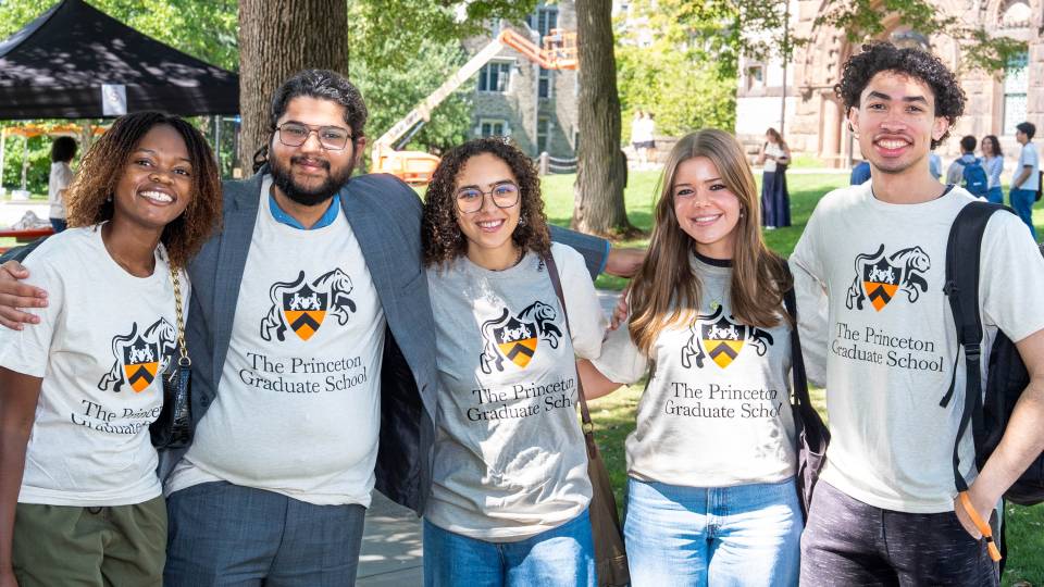 A group of smiling students in Graduate School t-shirts stand side-by-side on campus.
