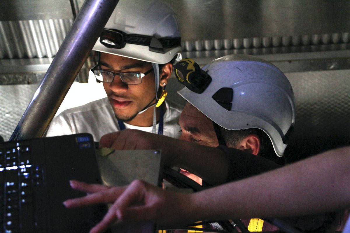 Two students wearing white hard hats look into the machinery of CERN