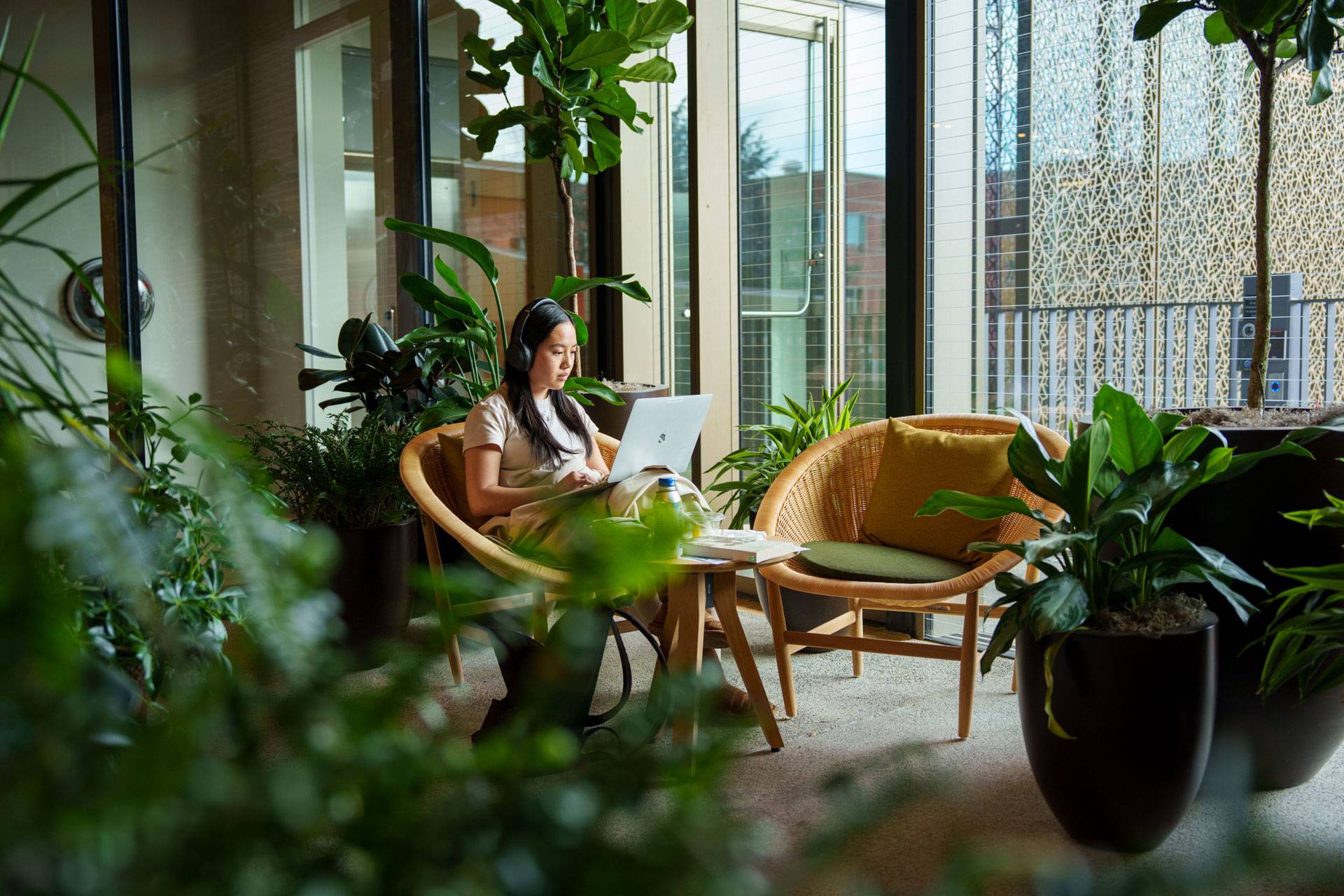 A student sits with headphones typing on a laptop in plant-filled room
