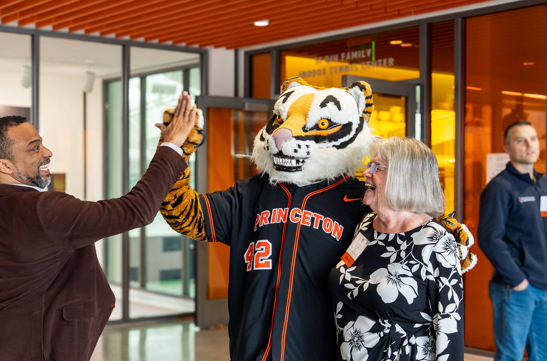 A man wearing a brown jacket is high-fiving the Princeton tiger mascot. A lady is smiling nearby.