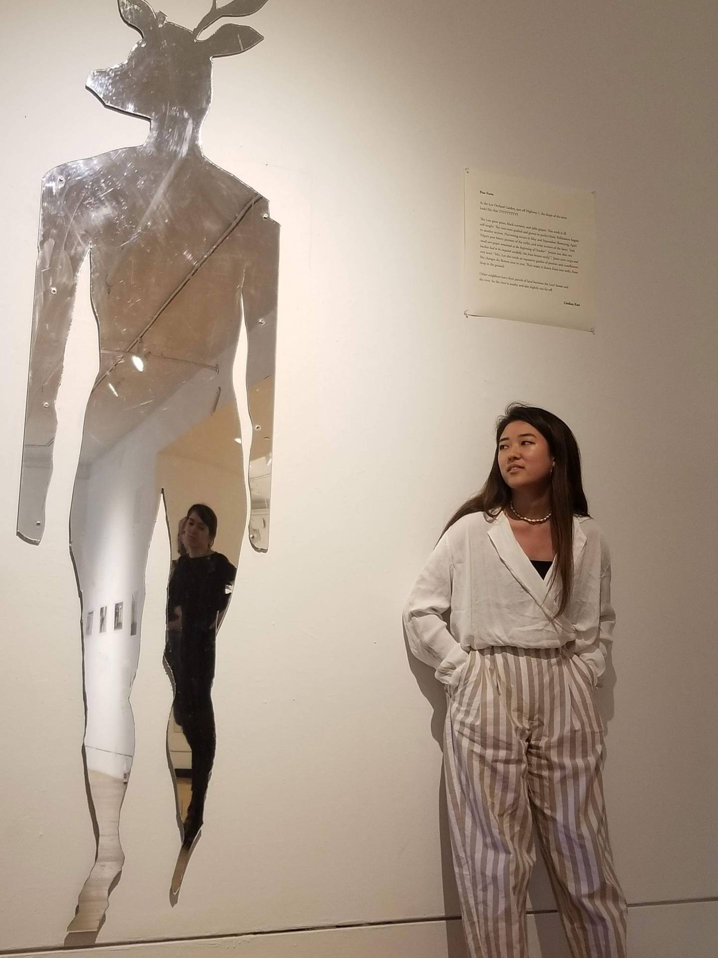 A student stands next to her art: a reflective deer-humanoid shape
