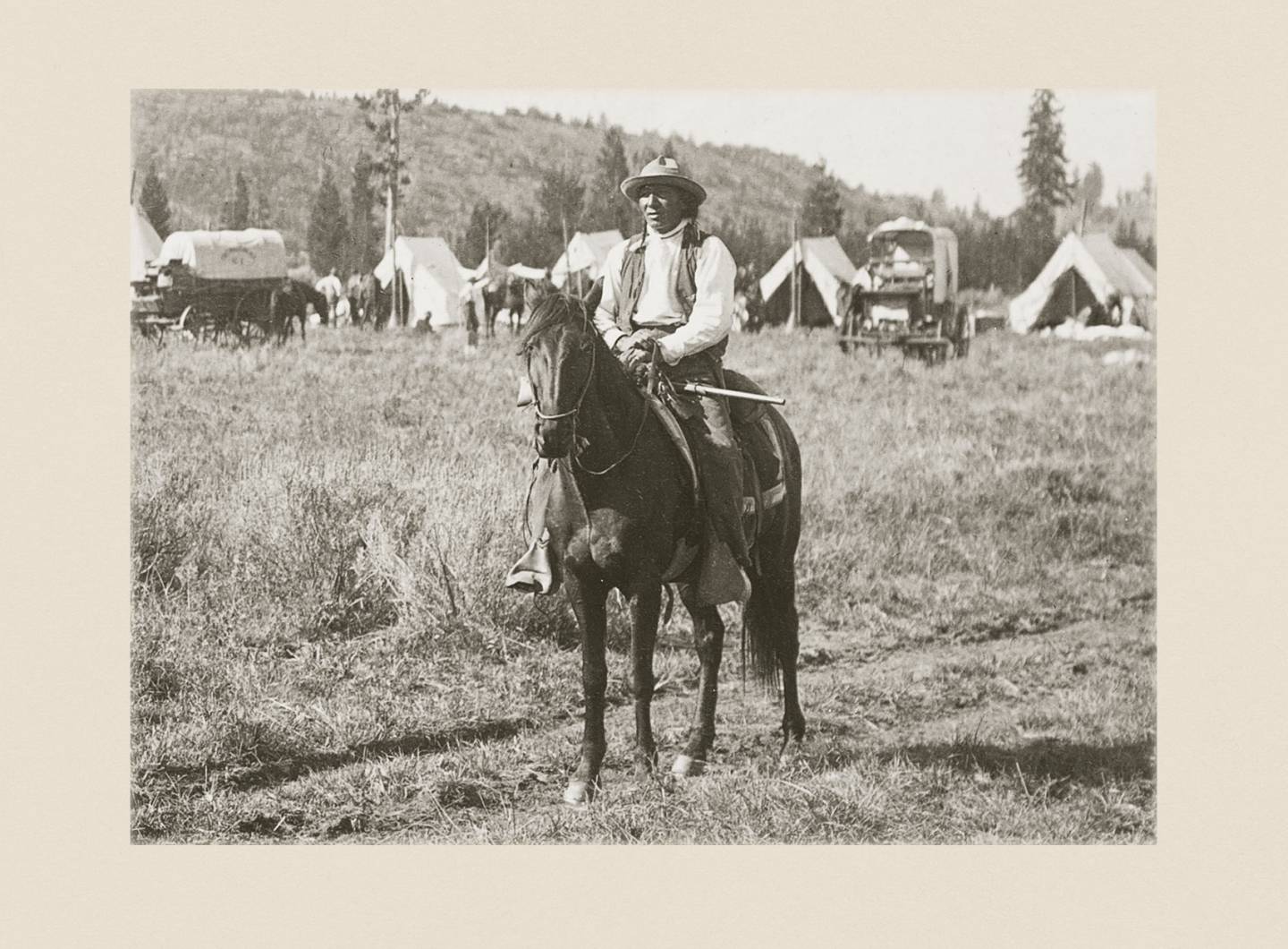 A man sits on a horse with a settlement of tents and mountains behind