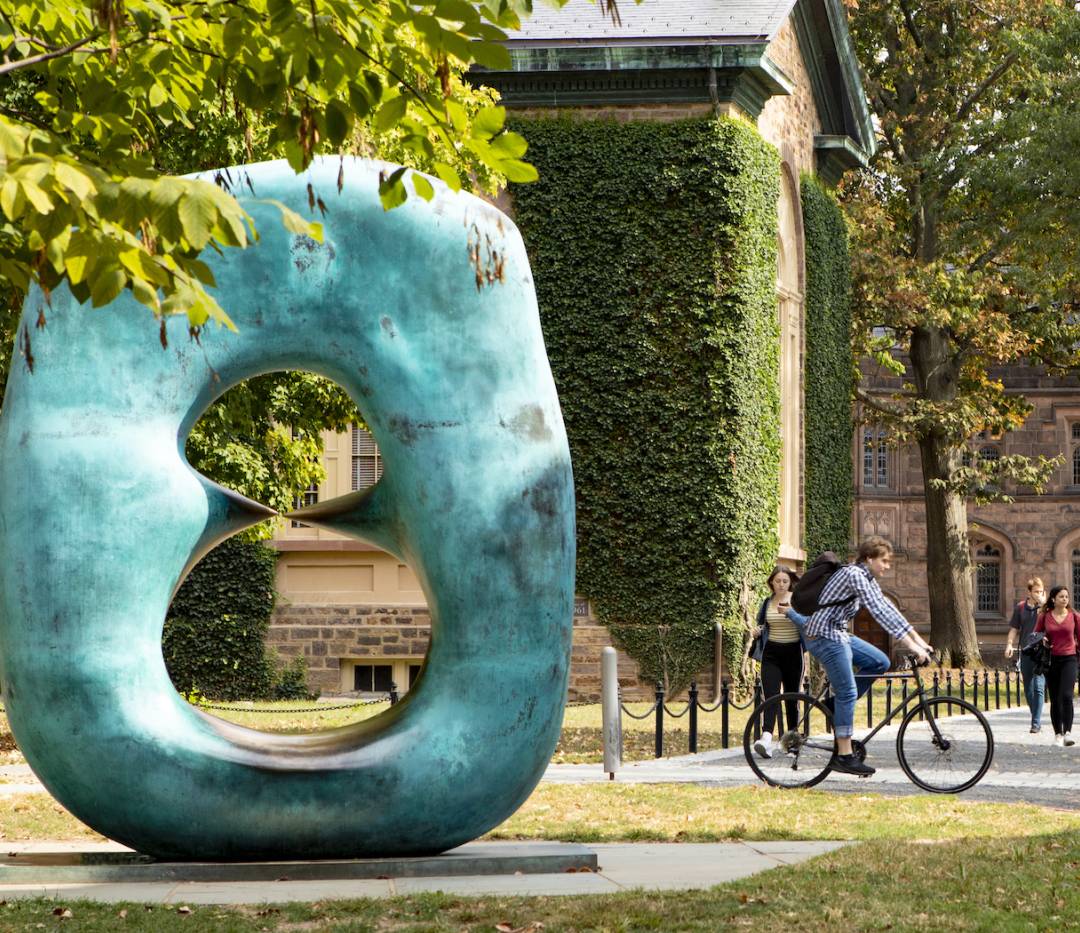 Student on bicycle riding past Oval with Points sculpture outside Nassau Hall near Cannon Green.