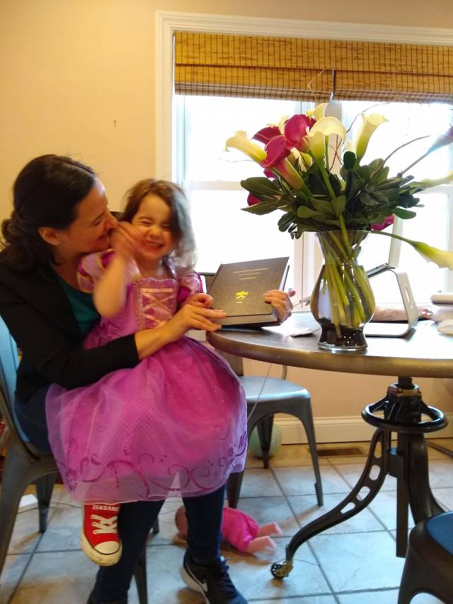 Hannah Rubinton sits at her laptop with a child in a pink tutu on her lap